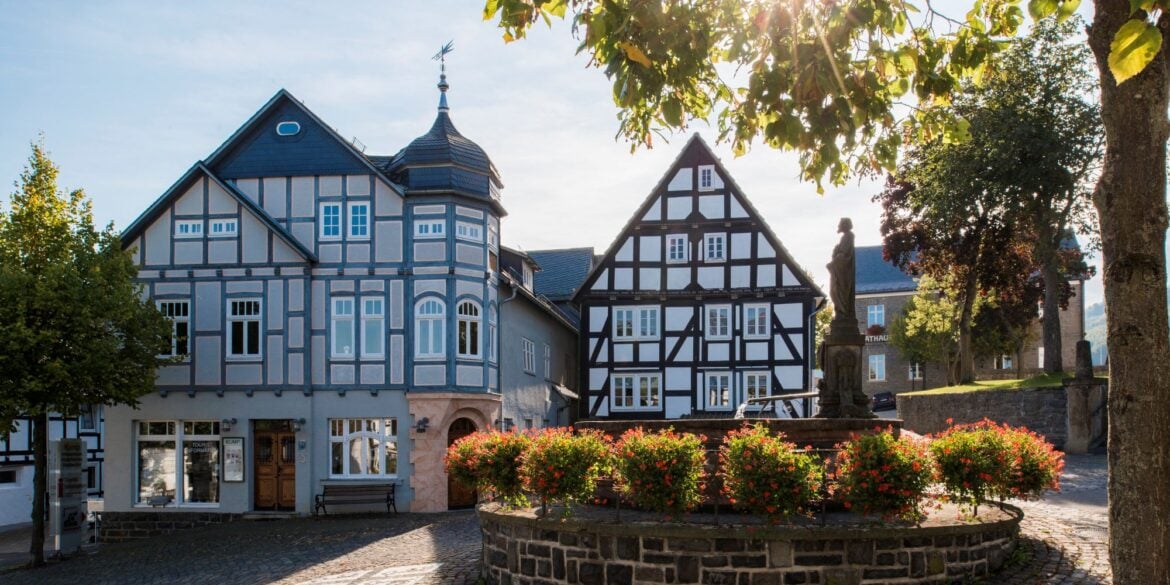 Zwei gut gepflegte Fachwerkhäuser stehen an einem gepflasterten Platz, davor ein gemauerter Brunnen mit Statue, um den kreisrund ein Blumenbeet angelegt wurde. Die Sonne scheint auf das Ensemble.