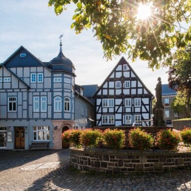 Zwei gut gepflegte Fachwerkhäuser stehen an einem gepflasterten Platz, davor ein gemauerter Brunnen mit Statue, um den kreisrund ein Blumenbeet angelegt wurde. Die Sonne scheint auf das Ensemble.