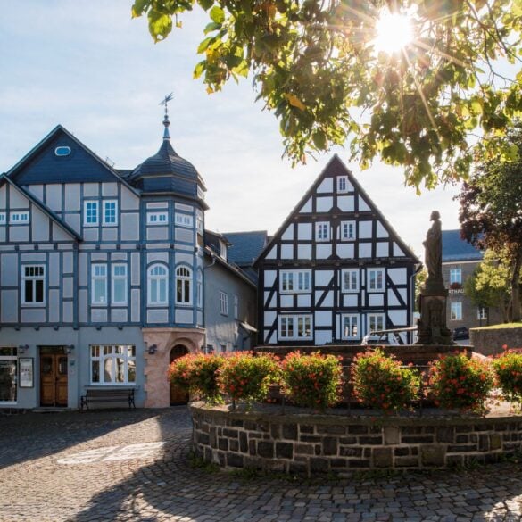 Zwei gut gepflegte Fachwerkhäuser stehen an einem gepflasterten Platz, davor ein gemauerter Brunnen mit Statue, um den kreisrund ein Blumenbeet angelegt wurde. Die Sonne scheint auf das Ensemble.
