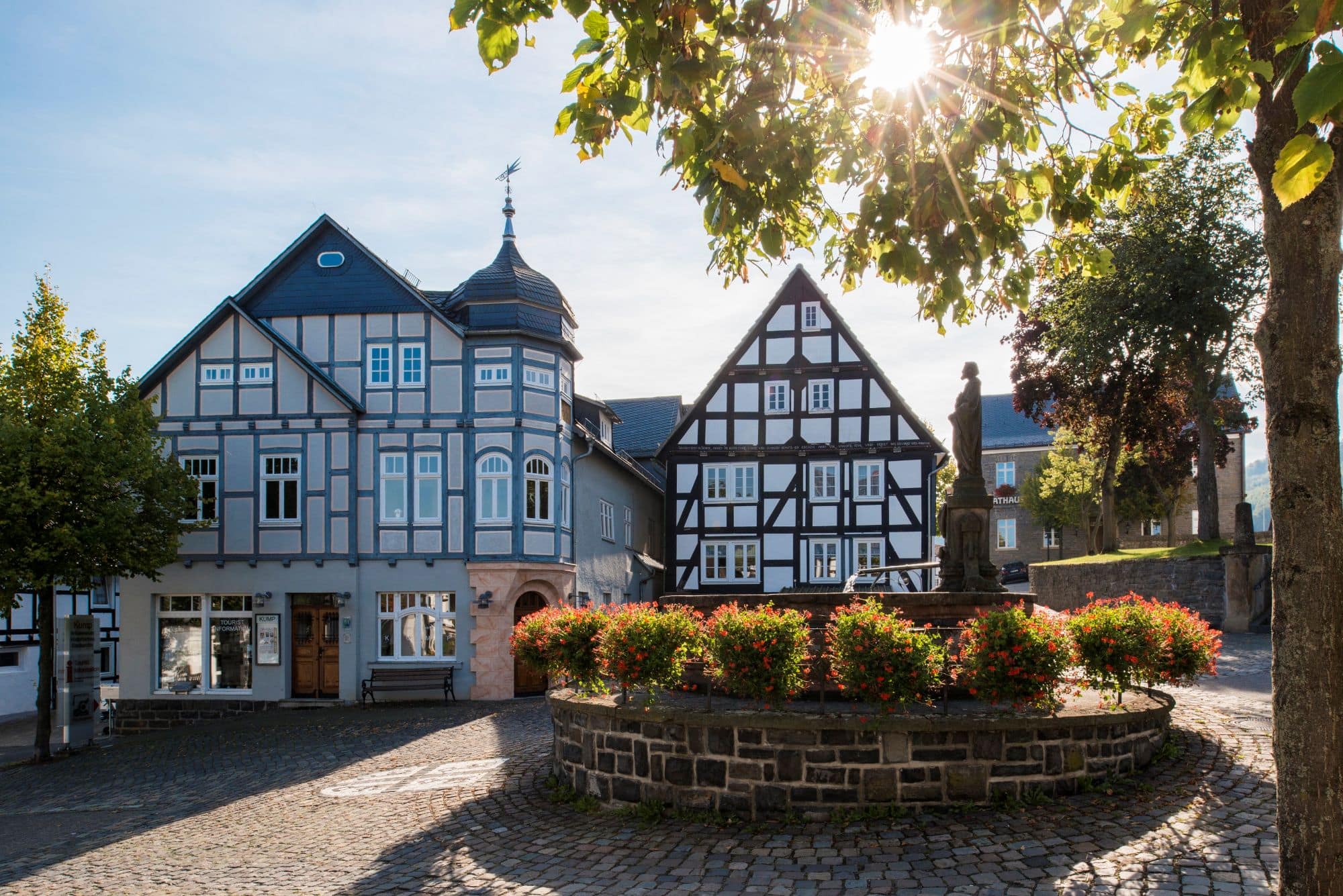 Zwei gut gepflegte Fachwerkhäuser stehen an einem gepflasterten Platz, davor ein gemauerter Brunnen mit Statue, um den kreisrund ein Blumenbeet angelegt wurde. Die Sonne scheint auf das Ensemble.