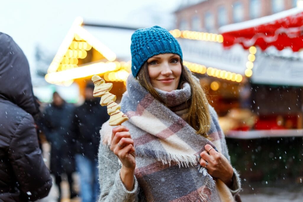 A woman in winter clothing at a christmas market, holding a sweet snack on a stick and smiling into the camera.
