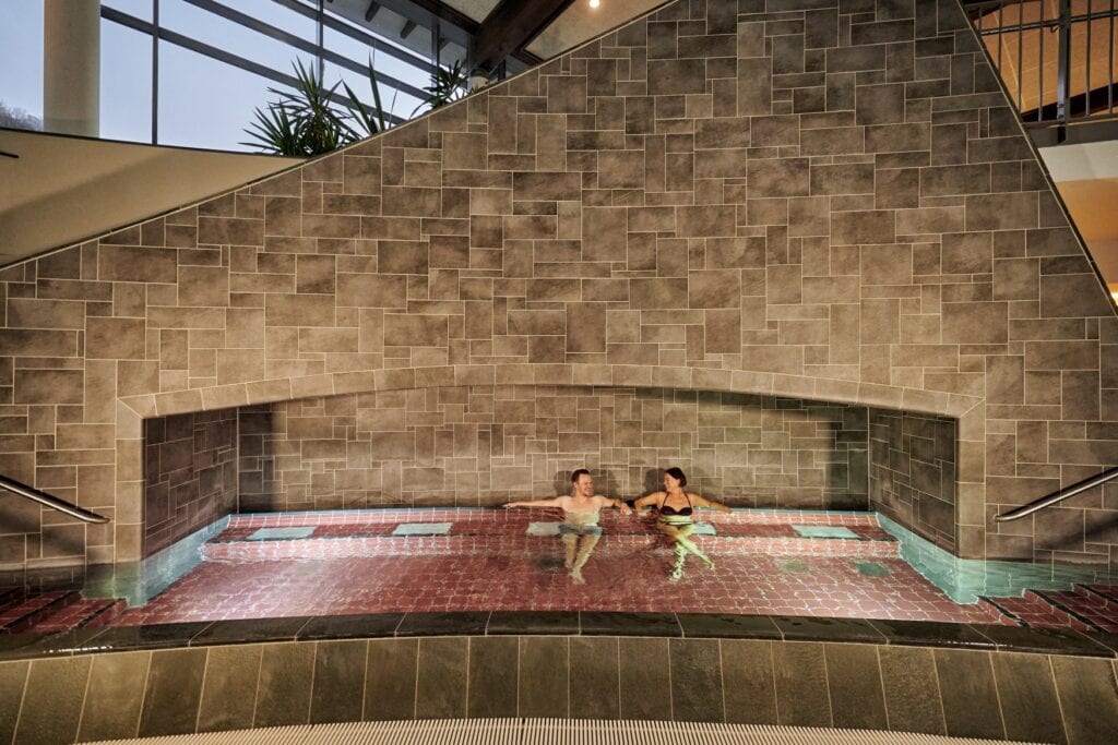 Zwei Personen liegen in einem geziegelten Flachwasserbecken in einer gemauerten Alkove in einer Therme und unterhalten sich.