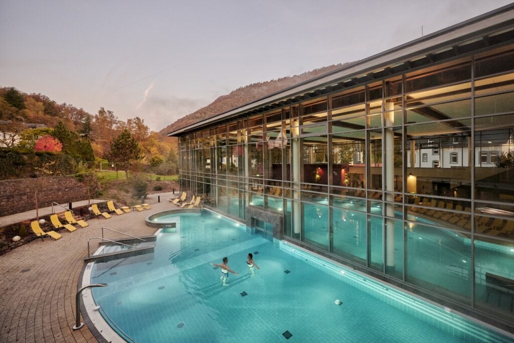 Drohnenaufnahme einer Therme von außen. Zwei Personen stehen in einem halbrunden Außenbecken, dass durch einen Durchbruch mit dem Innenbereich verbunden ist. Die gesamte Front der Therme ist verglast, innen ist das Becken ebenfalls halbrund angelegt. Auf der linken Bildseite ein grüner Hang mit herbstlichen Bäumen.