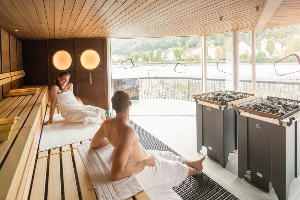 Ein Mann und eine Frau sitzen in einer Sauna und unterhalten sich. Links drei Sitzebenen übereinander, rechts öffnet eine breite Fensterfront den Blick auf einen Fluss und das gegenüberliegende Ufer.