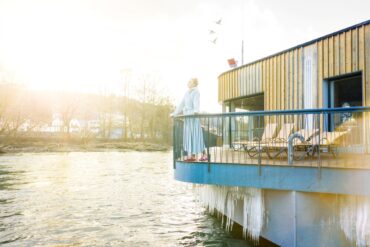 Eine Frau in Bademantel steht auf der Terrasse einer hölzernen Sauna über einem Fluss. Unter der Terrasse ragen mehrere Eiszapfen bis fast ins Wasser. Von links scheint kräftig die Sonne ins Bild.