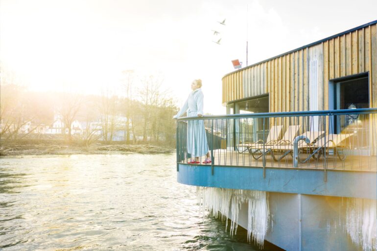 Eine Frau in Bademantel steht auf der Terrasse einer hölzernen Sauna über einem Fluss. Unter der Terrasse ragen mehrere Eiszapfen bis fast ins Wasser. Von links scheint kräftig die Sonne ins Bild.