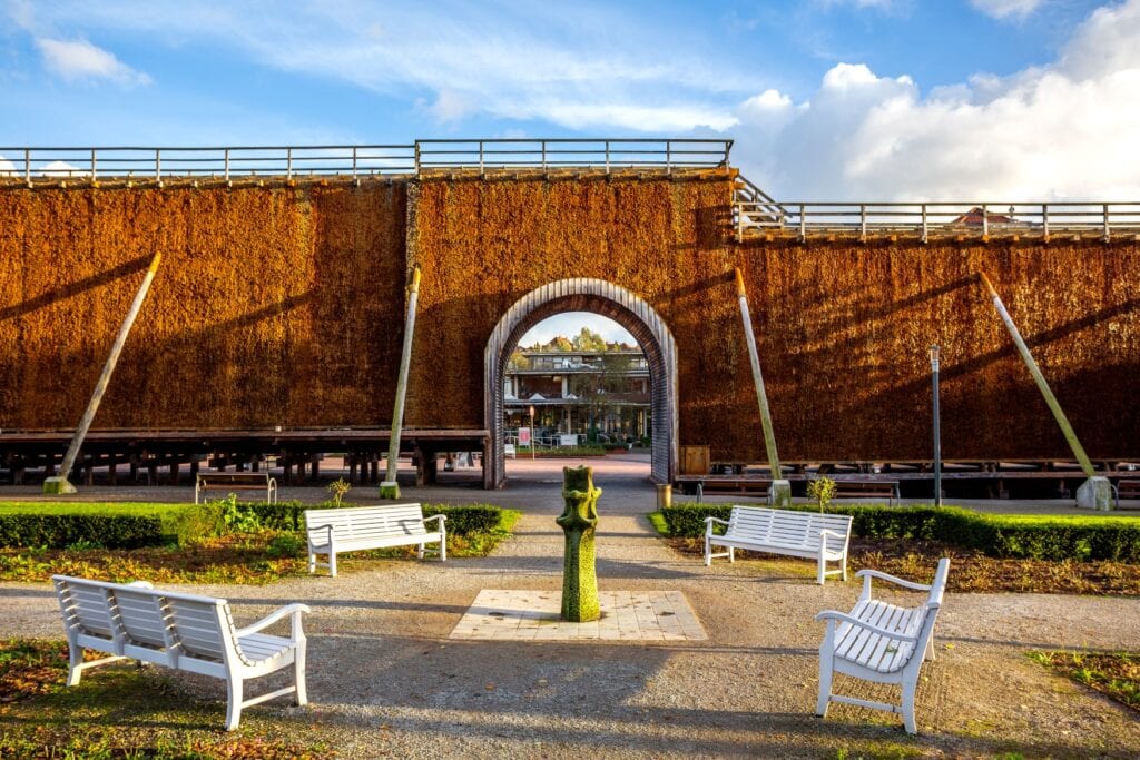 Ein rostrotes Gradierwerk mit einem mittigen Durchgang, genau von links nach rechts aufgenommen. Davor eine Sitzgruppe mit vier weißen Bänken.