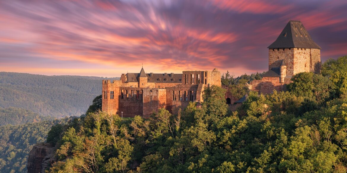 Auf einem Vorsprung über einem bewaldeten Tal thront eine gut erhaltene Burgruine aus rotem Sandstein mit einem mächtigen Eckturm. Hinter der Ruine geht brennend rot die Sonne unter.