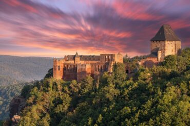 Auf einem Vorsprung über einem bewaldeten Tal thront eine gut erhaltene Burgruine aus rotem Sandstein mit einem mächtigen Eckturm. Hinter der Ruine geht brennend rot die Sonne unter.