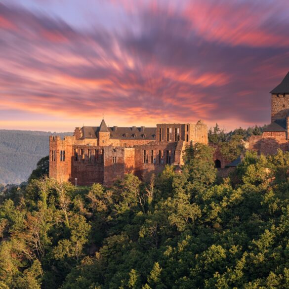 Auf einem Vorsprung über einem bewaldeten Tal thront eine gut erhaltene Burgruine aus rotem Sandstein mit einem mächtigen Eckturm. Hinter der Ruine geht brennend rot die Sonne unter.