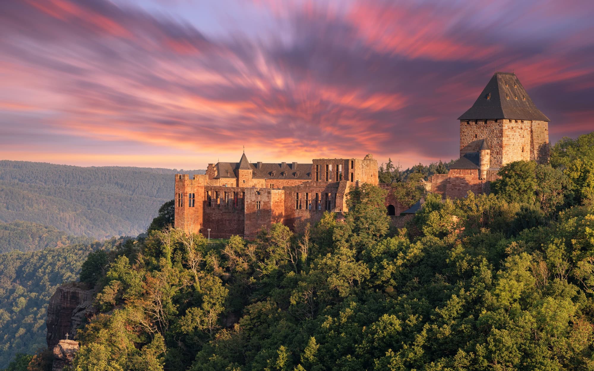 Auf einem Vorsprung über einem bewaldeten Tal thront eine gut erhaltene Burgruine aus rotem Sandstein mit einem mächtigen Eckturm. Hinter der Ruine geht brennend rot die Sonne unter.