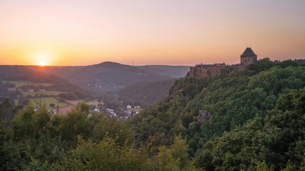 Über einem bewaldeten Tal mit einigen Siedlungen thront eine Burgruine mit einem massiven Eckturm aus roten Bundsandstein. Links geht am Horizont die Sonne unter.