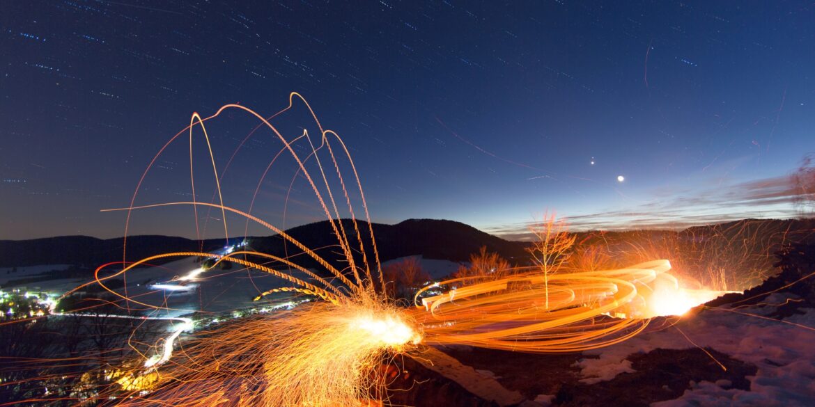 Langzeitaufnahme beim Scheibenschlagen in Bernau; Auf einem Hang sind die Lichtspuren eines Feuers zu sehen, eine Spur führt in einem hohen Bogen ins im Hintergrund liegende Tal. Darüber der Nachthimmel.