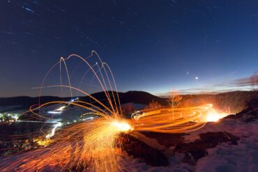 Langzeitaufnahme beim Scheibenschlagen in Bernau; Auf einem Hang sind die Lichtspuren eines Feuers zu sehen, eine Spur führt in einem hohen Bogen ins im Hintergrund liegende Tal. Darüber der Nachthimmel.