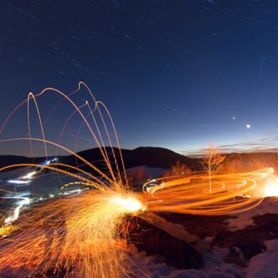 Langzeitaufnahme beim Scheibenschlagen in Bernau; Auf einem Hang sind die Lichtspuren eines Feuers zu sehen, eine Spur führt in einem hohen Bogen ins im Hintergrund liegende Tal. Darüber der Nachthimmel.
