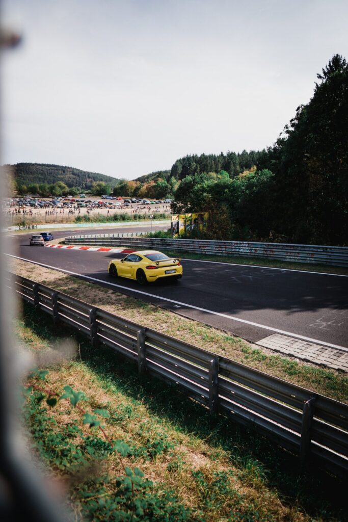 A racetrack photographed from the side of the road; a yellow Porsche approaches a curve. Numerous vehicles and spectators are standing in a pasture behind the racetrack in the background.