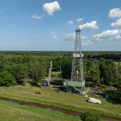 Auf einem grünen Feld steht ein gut 50 Meter hoher Erdölturm aus Stahl, der die hinter ihm stehenden Bäume weit überragt. Aufgenommen mit der Drohne.
