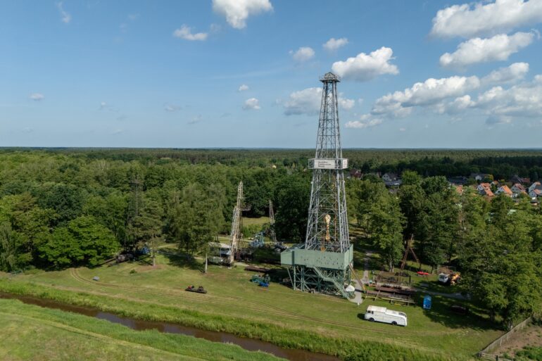 Auf einem grünen Feld steht ein gut 50 Meter hoher Erdölturm aus Stahl, der die hinter ihm stehenden Bäume weit überragt. Aufgenommen mit der Drohne.