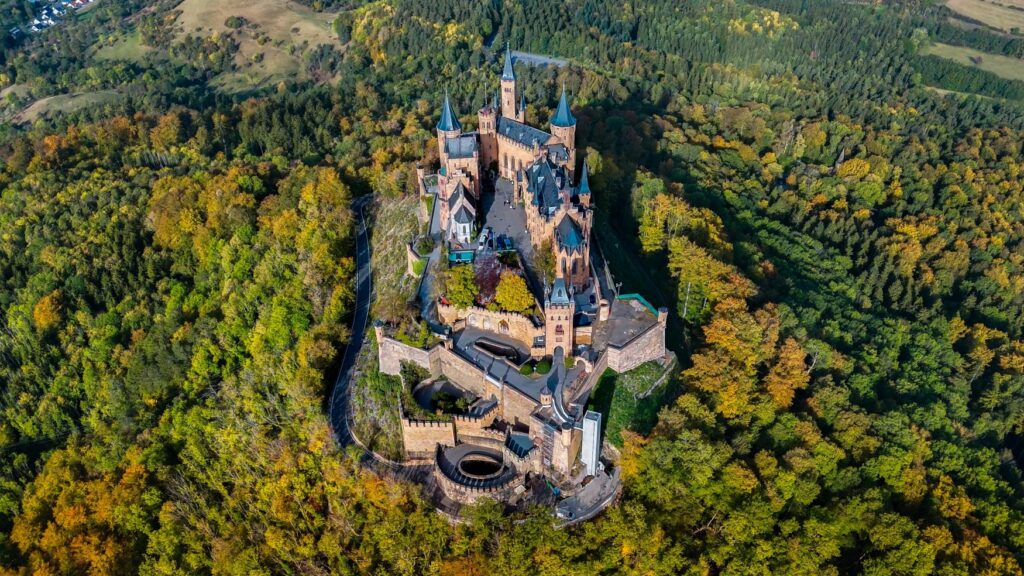 Drone shot of a hilltop castle on a wooded mountain. The castle stretches from top to bottom, and on the right-hand side, a church appears to be built into the castle. The courtyard is divided into several levels, and a road leads from the bottom around the castle towards the valley.