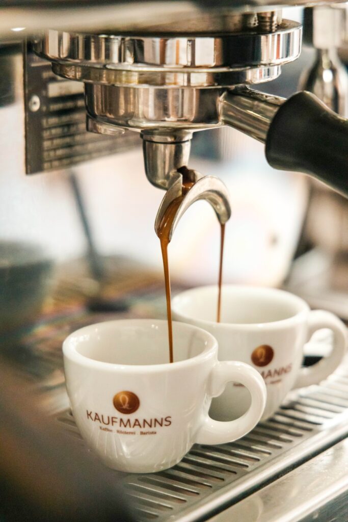 Close-up of a portafilter machine, from which espresso is currently being poured into two cups standing next to each other. The word “Kaufmanns” can be read on the cups.
