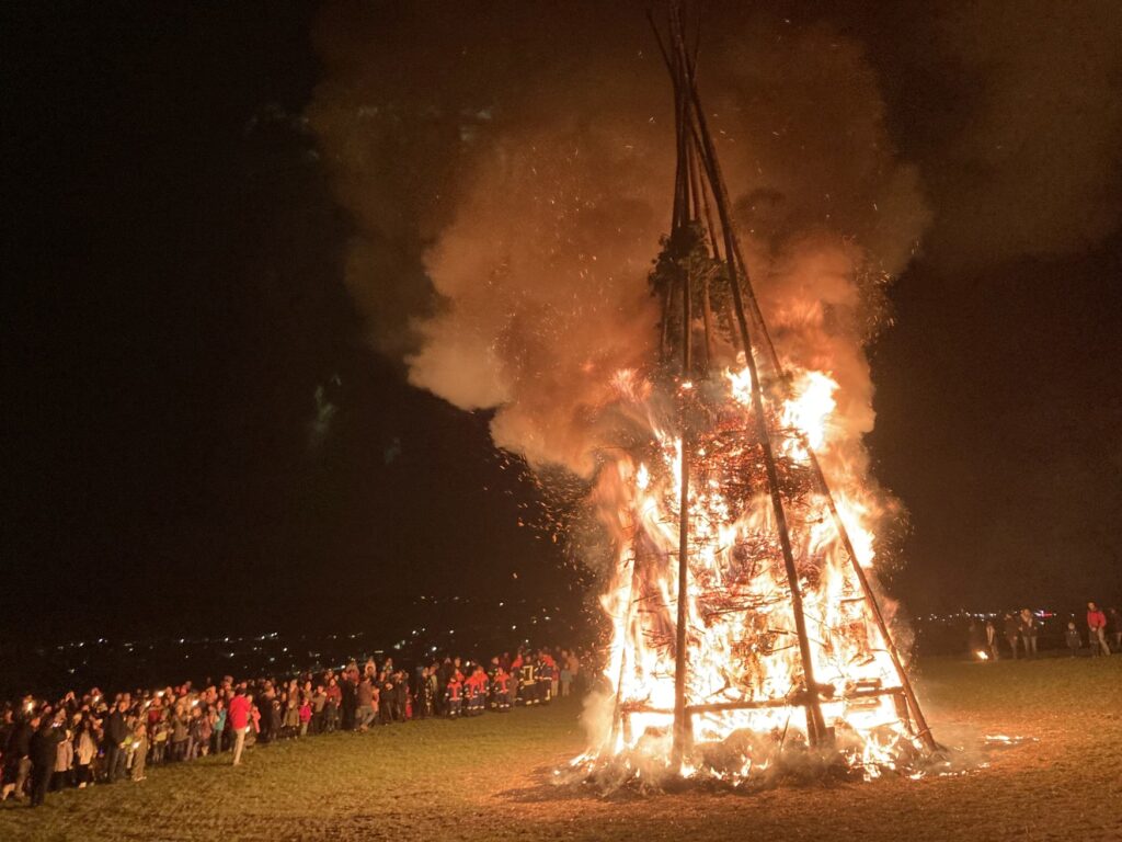 Ein mehrere Meter hoch aufgeschichtetes Feuer brennt in der dunklen Nacht. In einem Halbkreis darum steht eine große Menschenmenge.