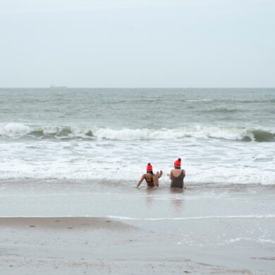 Zwei Frauen in Schwimmkleidung und mit orangenen Mützen sitzen in der Brandung der winterlichen Nordsee.