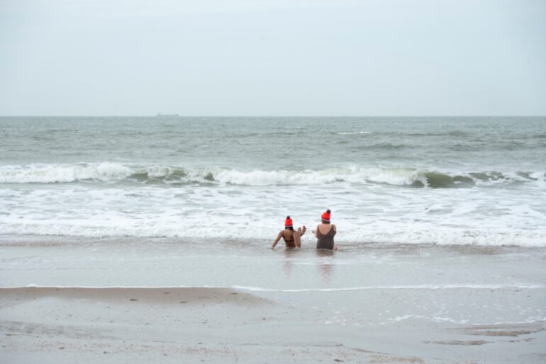 Zwei Frauen in Schwimmkleidung und mit orangenen Mützen sitzen in der Brandung der winterlichen Nordsee.