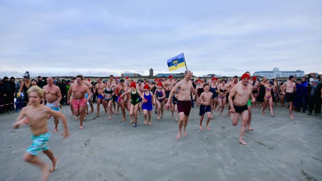 Zahlreiche Menschen in Schwimmkleidung rennen durch flaches Wasser an einem Strand in Richtung des Betrachters.