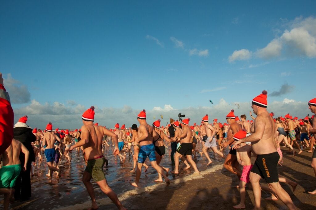 Zahlreiche Menschen in Schwimmkleidung und mit roten Mützen auf dem Kopf rennen über einen Strand in Richtung Meer.