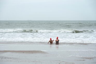 Two women in swimsuits and orange hats sit in the surf of the wintry North Sea.