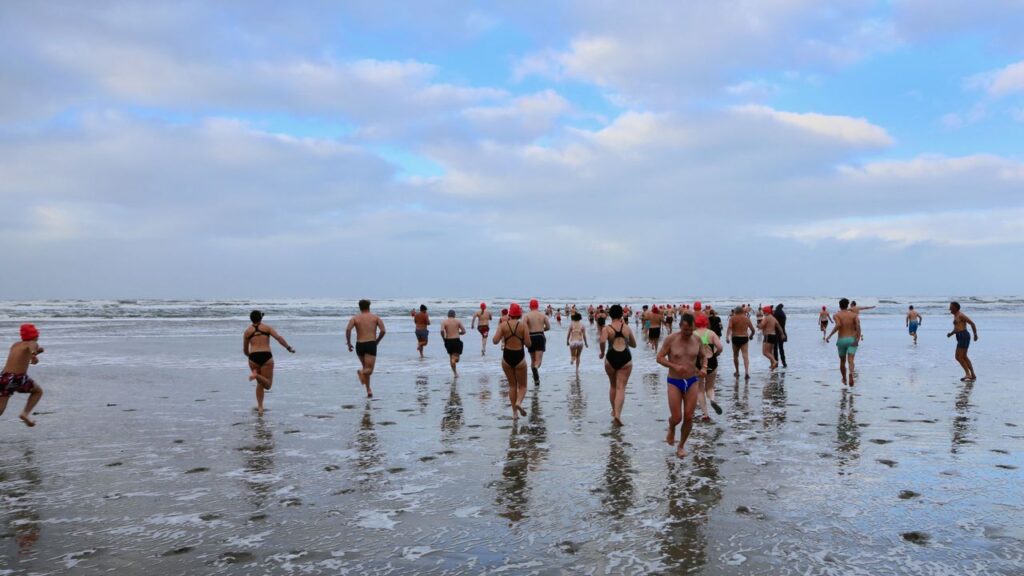 Several people in swimwear are running across a wet beach toward the sea.