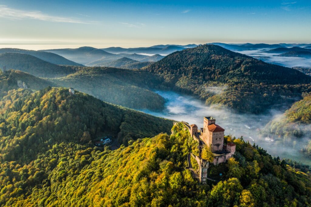 Drone shot of a hilltop castle made of red sandstone, rising from the forest at the bottom of the picture. Surrounded by the hilly Palatinate Forest, with fog hanging in its valleys.