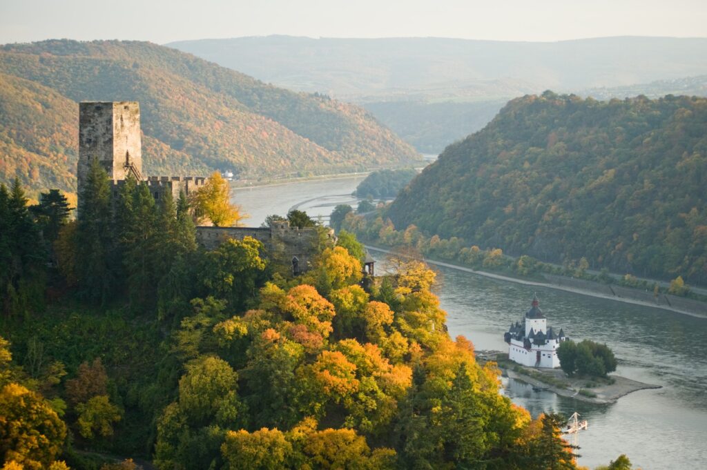 View of the Middle Rhine Valley from a hilltop. On the left, castle ruins rise above the autumnal forest; on the right, a white customs castle stands on an island in the middle of the Rhine. In the background, the Rhine first curves to the right and then, barely visible, to the left again.