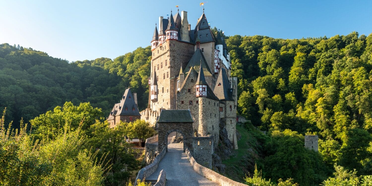 On a small hill in a wooded valley stands a narrow, very well-preserved castle. On the upper floors, the gray stone transitions into red and white half-timbering, and the roof is covered with gray slate. The castle can only be reached via a stone bridge. From the left, the sun shines brightly into the valley and onto the castle.