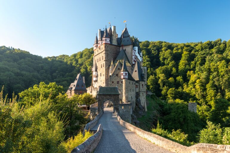 On a small hill in a wooded valley stands a narrow, very well-preserved castle. On the upper floors, the gray stone transitions into red and white half-timbering, and the roof is covered with gray slate. The castle can only be reached via a stone bridge. From the left, the sun shines brightly into the valley and onto the castle.