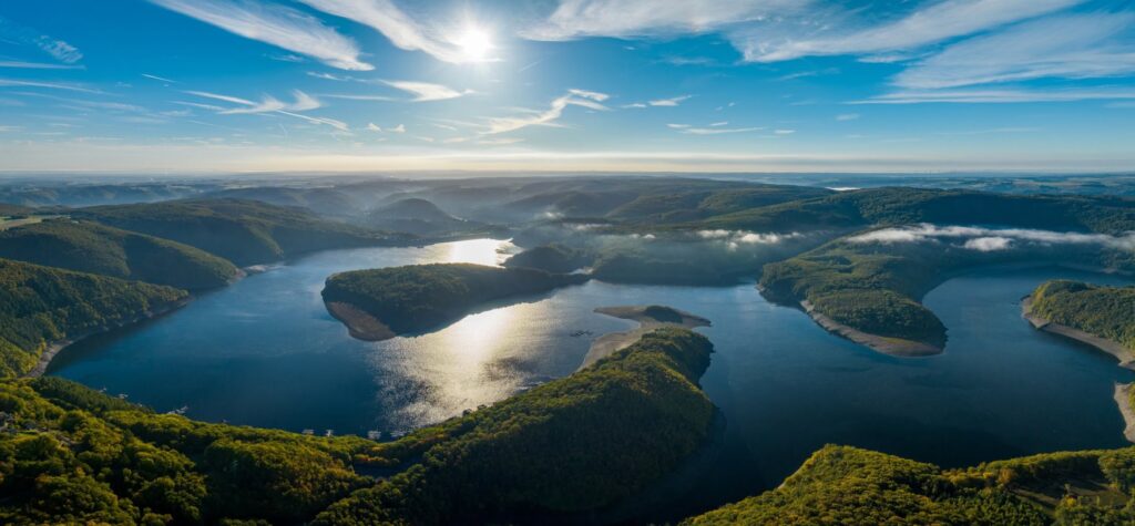 A large lake with several headlands jutting into it, photographed from the air. The land around the lake is covered with dense forest and is hilly, with some fog still lingering on the lake and in the valleys. The sky above is bright blue, and the sun is reflected on the water's surface.
