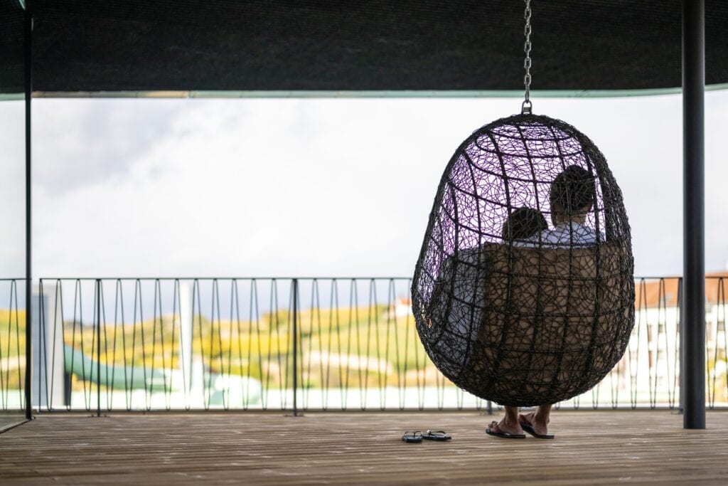 Zwei Personen sitzen auf einer überdachten Terrasse in einem hölzernen Hängekorb und blicken auf eine grüne, sonnenbeschienene Landschaft.