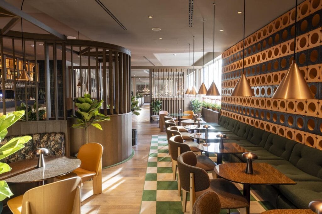 Lounge in einem Hotel; rechts eine lange, grüne Polsterbank, entlang der halbrunde Holztische aufgestellt sind, jeweils ein brauner Polsterstuhl gegenüber; links eine weitere Sitzecke, mit mit einer Polsterbank mit grünem Blumenmuster. Ein halbdurchlässiger, hölzerner Raumtrenner gewährt den Blick auf einen Barbereich.