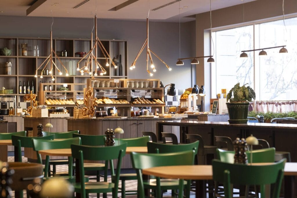 Ein Restaurant in einem modernen Landhausstil; viele kleine Holztische mit jeweils zwei tiefgrün gestrichenen Holzstühlen stehen im Vordergrund, dahinter eine Buffetbereich, auf dem ein Hotelfrühstück angerichtet ist.