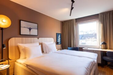 Double bed with white covers in a hotel room; the wall behind the bed is painted light brown, with two small pictures hanging on it. To the right of the bed, a large window lets in light. There is a narrow desk by the window, with two blue upholstered chairs to the left and right of it.