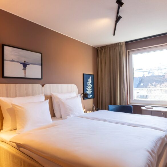 Double bed with white covers in a hotel room; the wall behind the bed is painted light brown, with two small pictures hanging on it. To the right of the bed, a large window lets in light. There is a narrow desk by the window, with two blue upholstered chairs to the left and right of it.