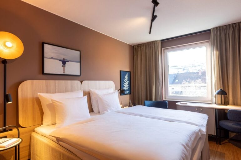 Double bed with white covers in a hotel room; the wall behind the bed is painted light brown, with two small pictures hanging on it. To the right of the bed, a large window lets in light. There is a narrow desk by the window, with two blue upholstered chairs to the left and right of it.