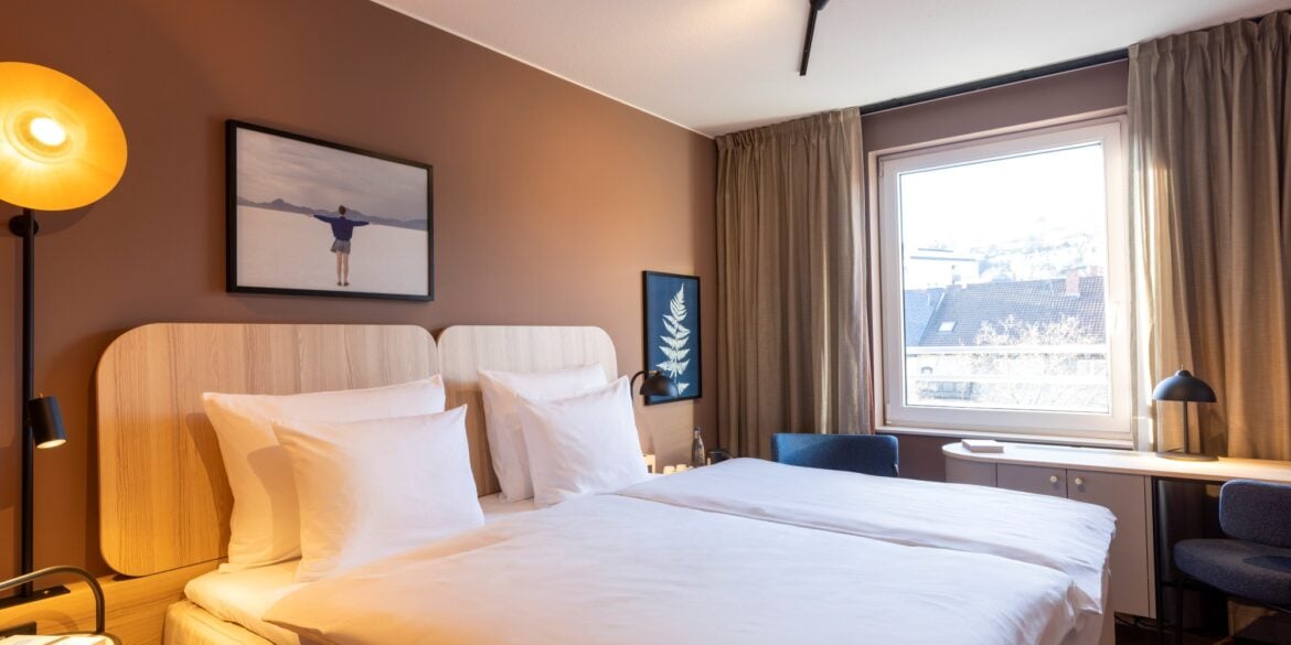 Doppelbett mit weißen Bezügen in einem Hotelzimmer; die Wand hinter dem Bett ist hellbraun gestrichen, zwei kleine Bilder hängen daran. Rechts vom Bett lässt ein großes Fenster Licht herein. Am Fenster steht eine schmaler Schreibtisch, links und rechts daneben zwei blaue Polsterstühle.