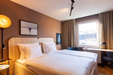 Doppelbett mit weißen Bezügen in einem Hotelzimmer; die Wand hinter dem Bett ist hellbraun gestrichen, zwei kleine Bilder hängen daran. Rechts vom Bett lässt ein großes Fenster Licht herein. Am Fenster steht eine schmaler Schreibtisch, links und rechts daneben zwei blaue Polsterstühle.