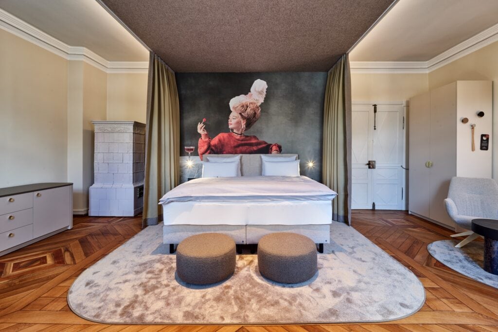 Große Suite in einem Hotel; genau mittig ein großes Doppelbett, dahinter ein Wandbild einer jungen Frau in einem roten, historischen Kleid mit weißer Turmperücke. Der Boden des Altbauzimmers ist aus Parkett, links vom Bett steht in der Ecke ein weißer Kachelofen, rechts ist eine hohe Doppeltür eingelassen.