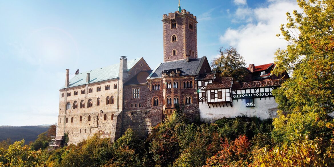 View of a hilltop castle stretching from the right edge of the picture to almost the left. The right part of the wall is covered by a half-timbered battlement; behind the central part made of red brick, a tall, angular tower rises up. The left part of the building is the tallest, with at least three stories and a green copper roof. The sky is bright blue, and the trees below the castle are already showing autumn colors.