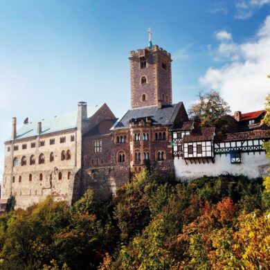 View of a hilltop castle stretching from the right edge of the picture to almost the left. The right part of the wall is covered by a half-timbered battlement; behind the central part made of red brick, a tall, angular tower rises up. The left part of the building is the tallest, with at least three stories and a green copper roof. The sky is bright blue, and the trees below the castle are already showing autumn colors.
