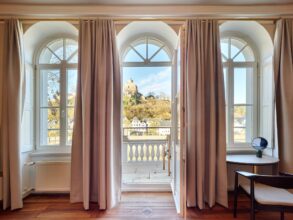 Zwei hohe Fenster und eine mittige Tür trennen ein Altbauzimmer mit Parkettboden von einem weoßen Balkon. Der Blick geht auf eine Burgruine in einem Wald.