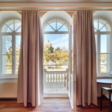 Zwei hohe Fenster und eine mittige Tür trennen ein Altbauzimmer mit Parkettboden von einem weoßen Balkon. Der Blick geht auf eine Burgruine in einem Wald.