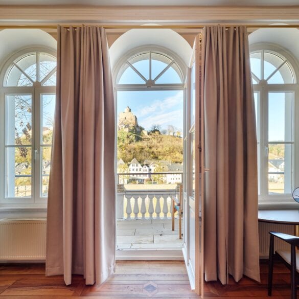 Zwei hohe Fenster und eine mittige Tür trennen ein Altbauzimmer mit Parkettboden von einem weoßen Balkon. Der Blick geht auf eine Burgruine in einem Wald.
